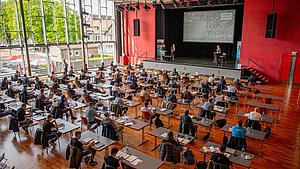 Sitzungssaal mit sitzenden Zuhörern, die auf den Redner blicken; Redner erläutert anhand von Darstellungen auf Großbildschirm